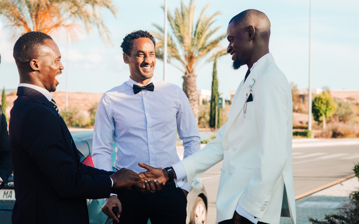 Trois amis en tenue de mariage rient ensemble, l'un d'eux serre la main d'un autre.